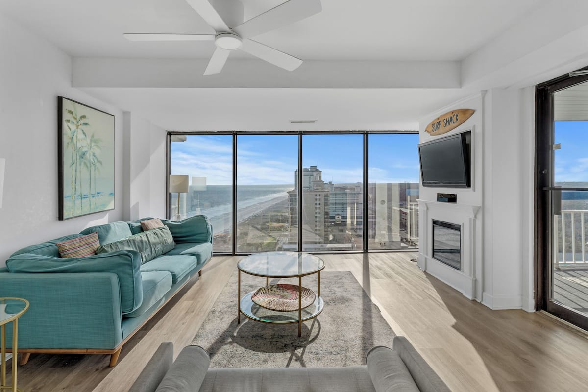 A modern living room with a teal sofa, a glass coffee table, and a view of the beach through large windows.