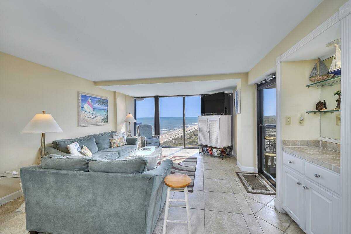 A living room with a grey sofa, a coffee table, a TV, and a view of the ocean through large windows.