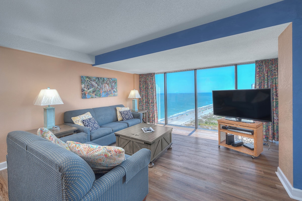 A living room with a large flat-screen TV, a sofa, a coffee table, and a view of the ocean.