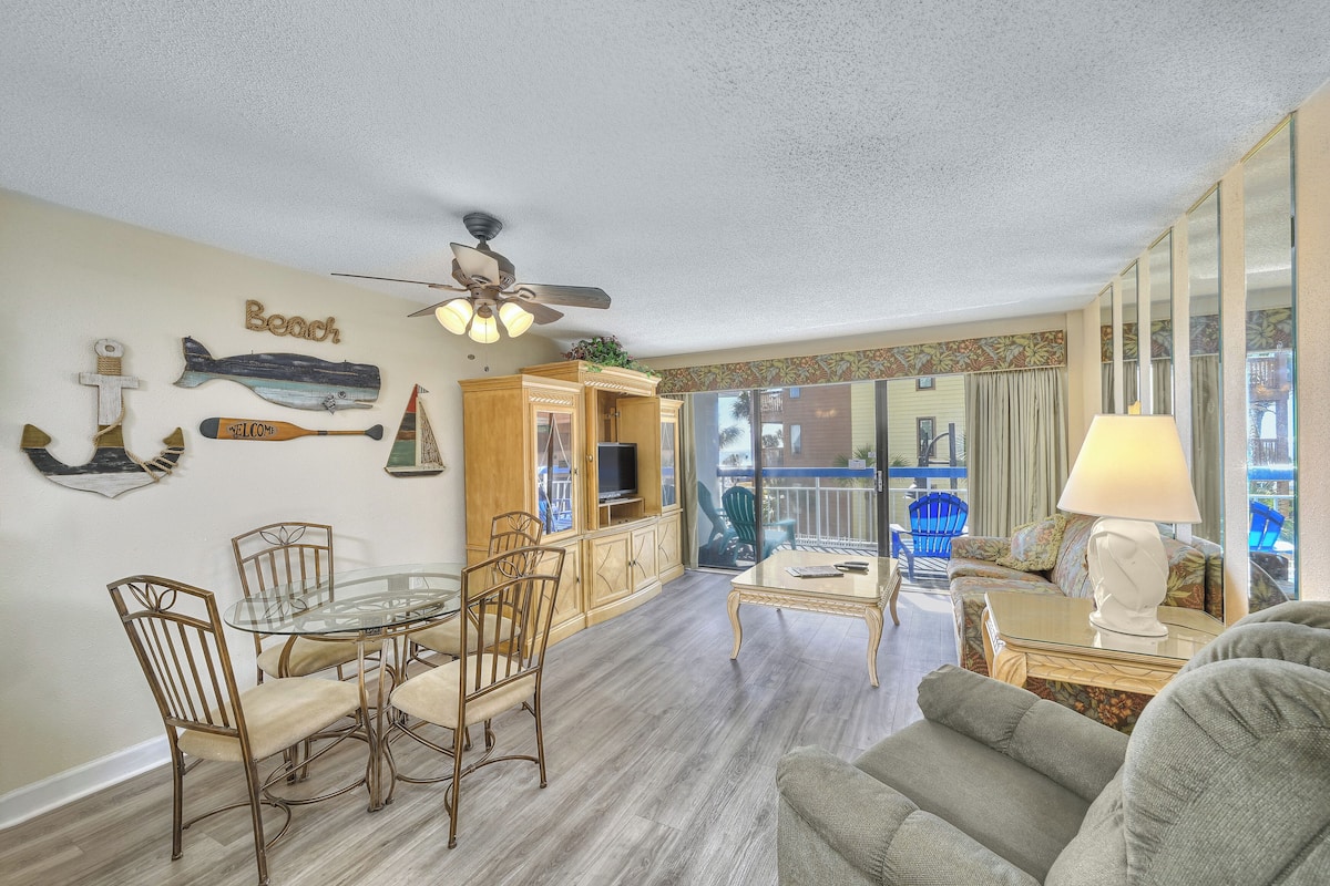 A spacious living room with a glass dining table, comfortable seating, a ceiling fan, and a view of the outdoors.