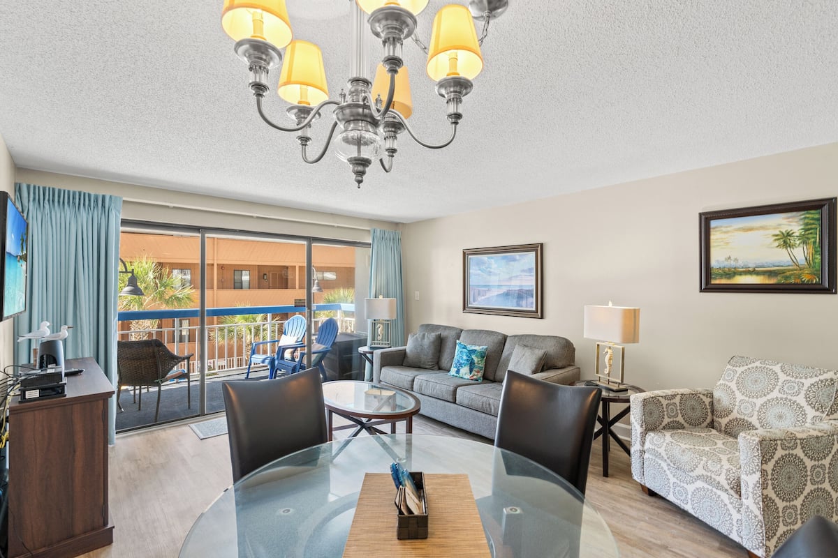 A spacious living room with a glass dining table, a sofa, and a patterned armchair. A chandelier hangs from the ceiling, and there is a balcony with a view of the outside.