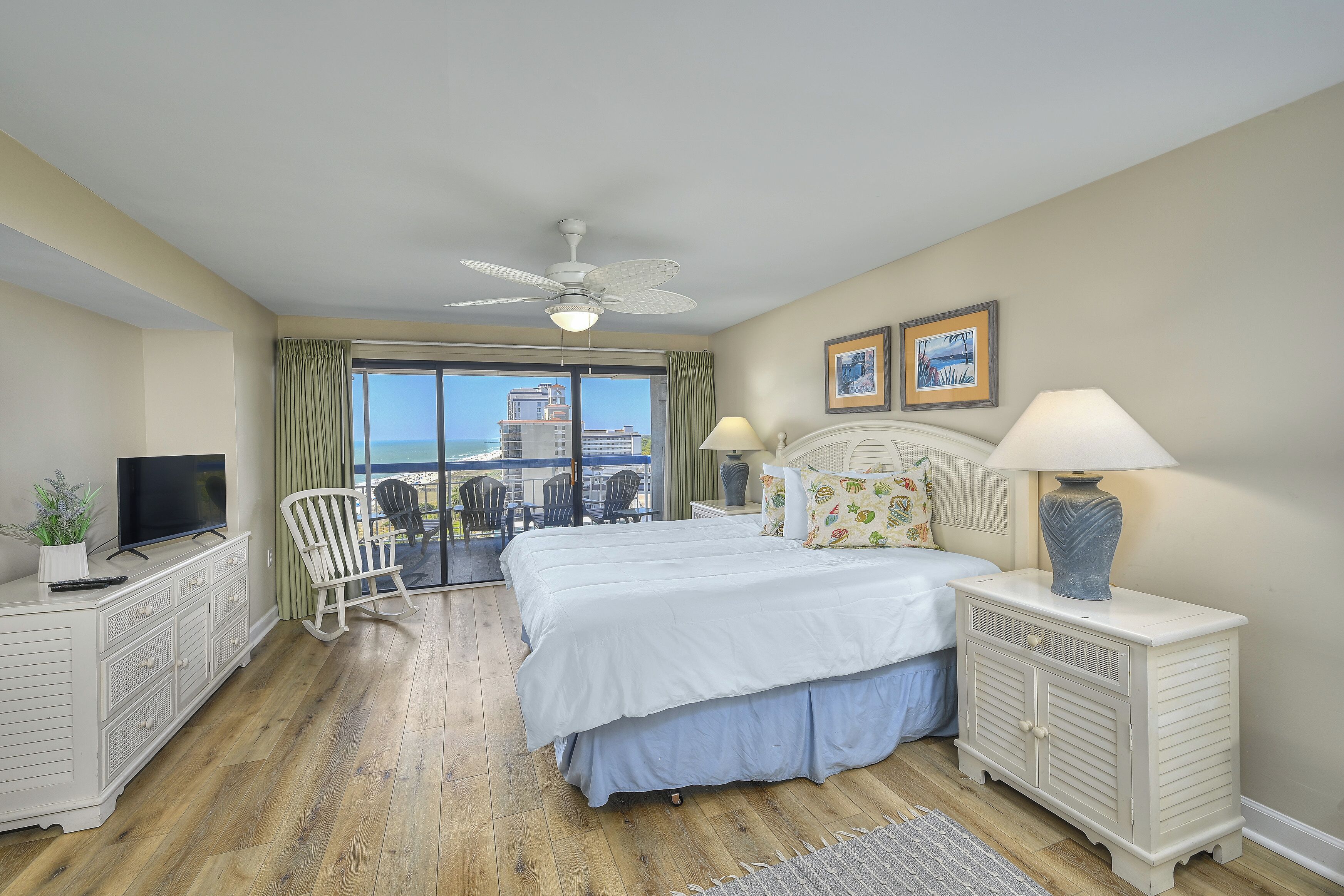 A bedroom with a bed, a TV, a ceiling fan, a nightstand, and a view of the ocean.