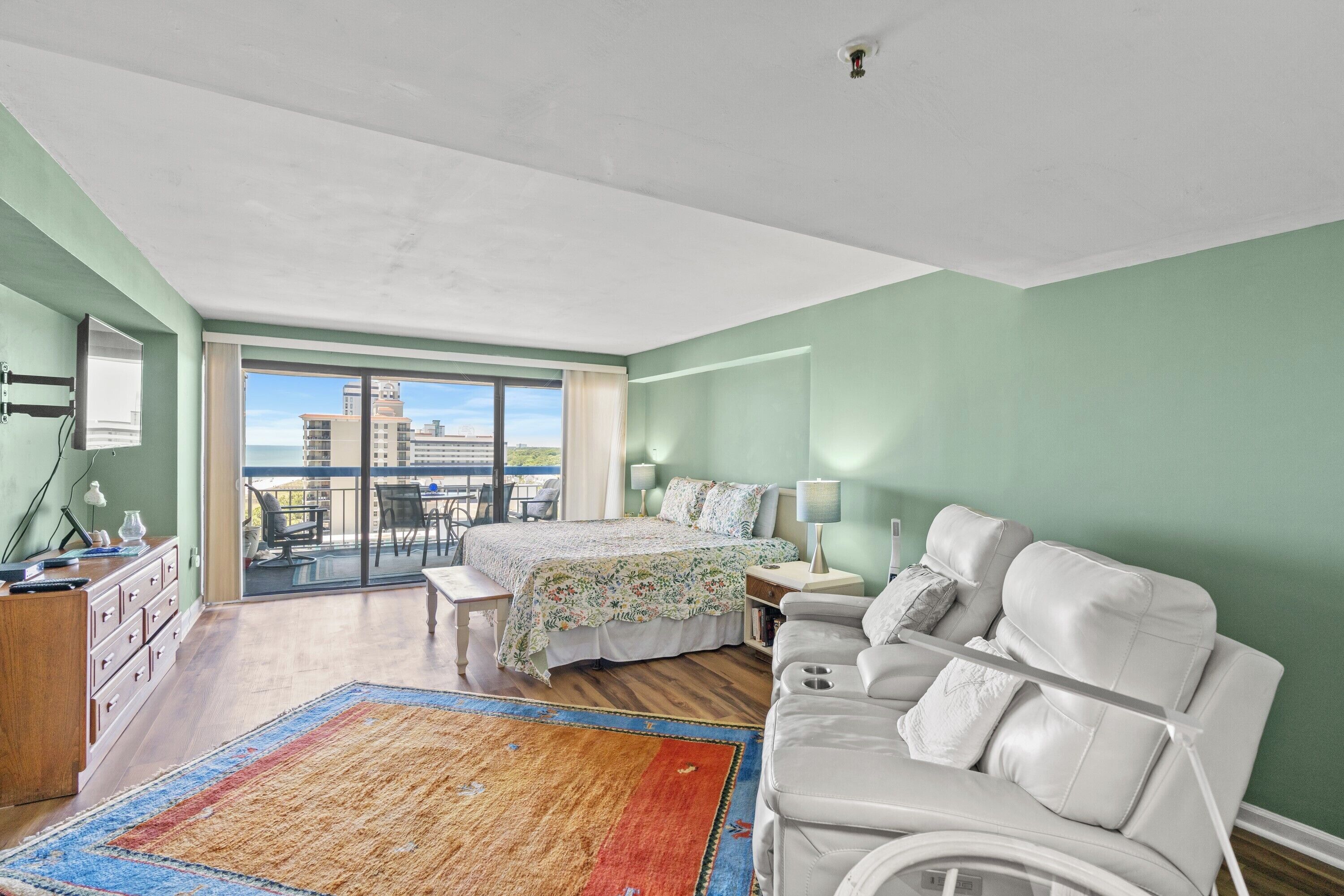 A bedroom with a bed, a white sofa, a desk, and a view of the city through a balcony.