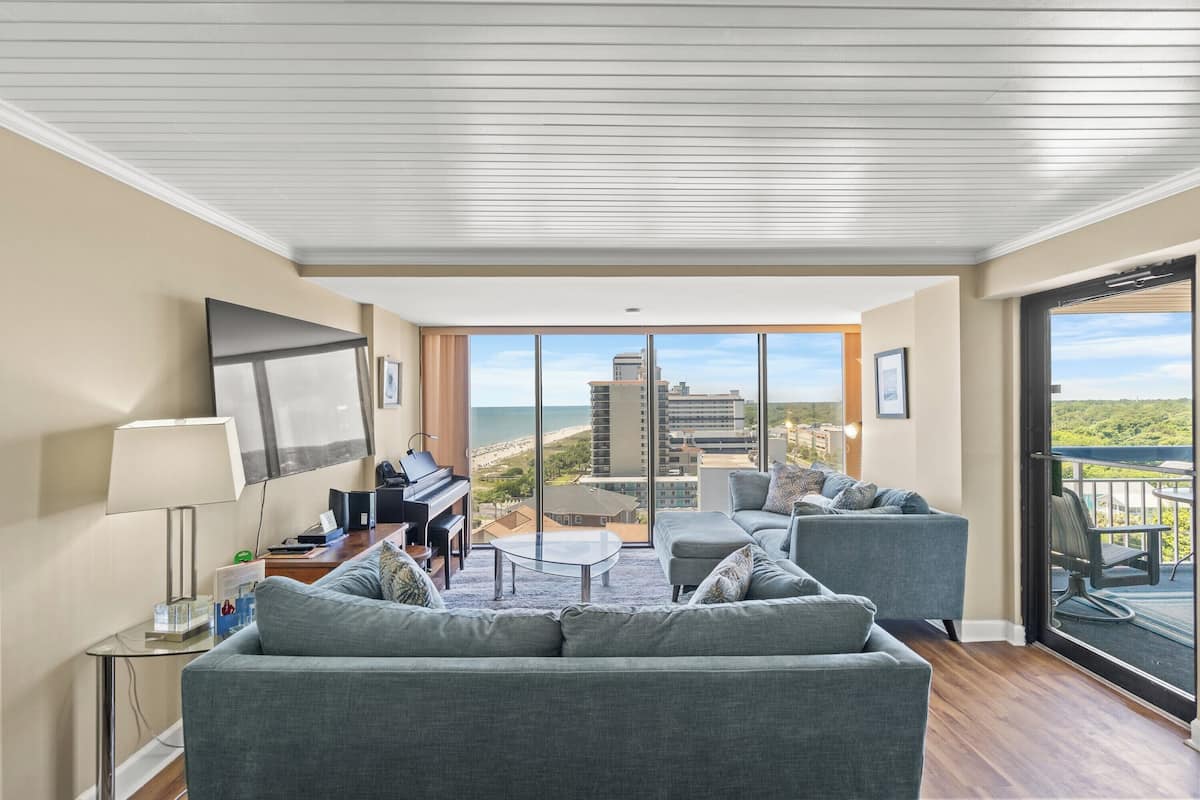 A modern living room with a piano, a sofa, a coffee table, and a view of the ocean through large windows.