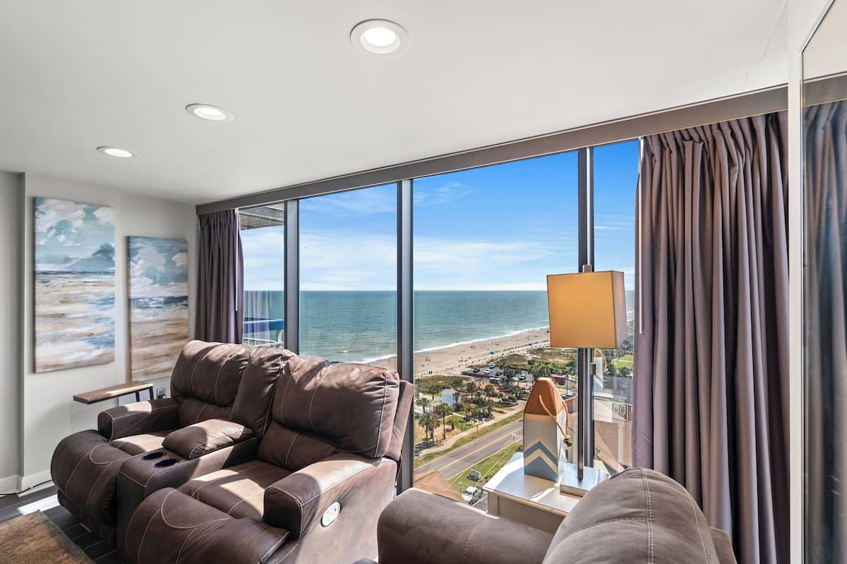 A modern living room with a reclining sofa, a large window with a view of the beach, and a lamp on a side table.