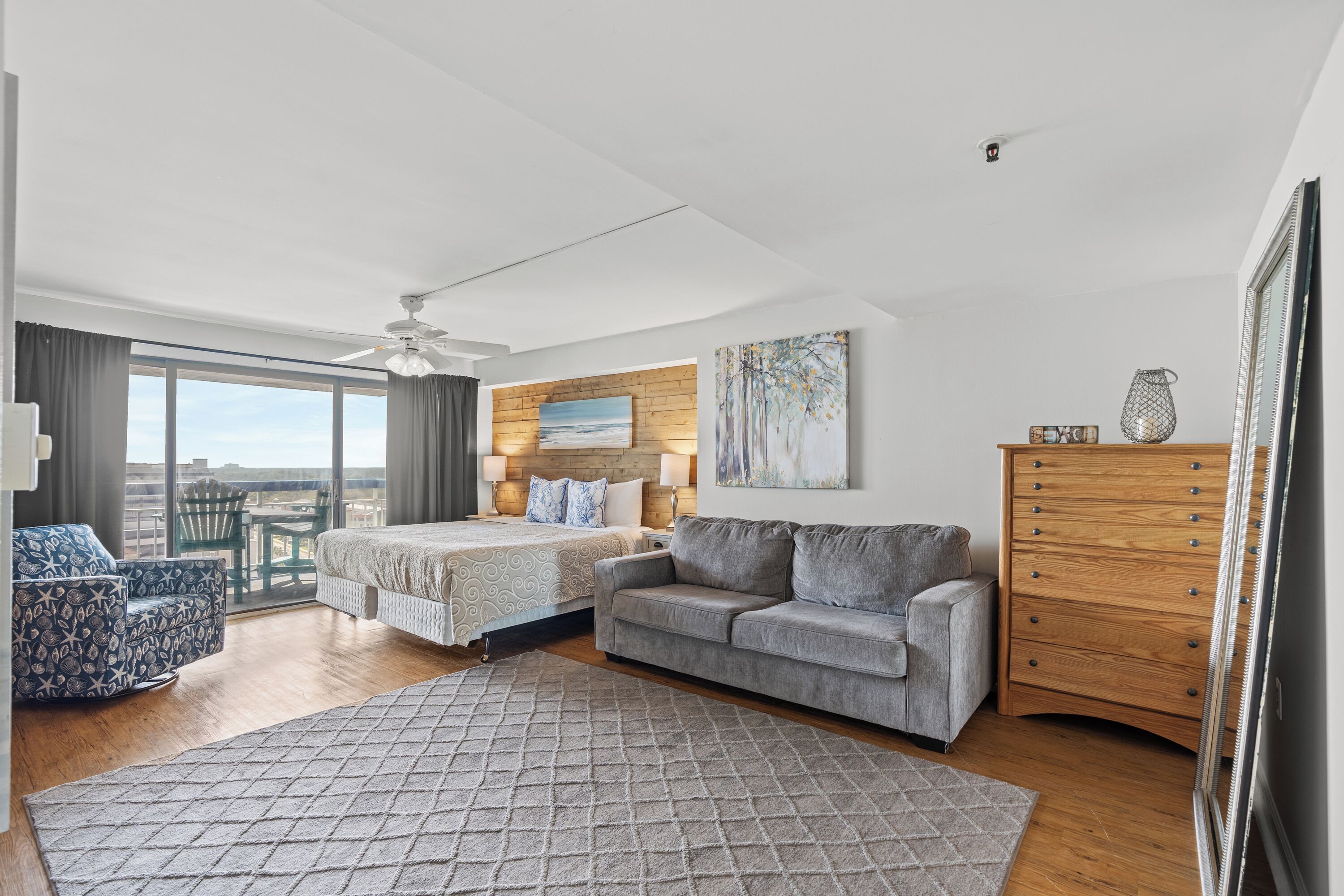 Une chambre moderne comprenant un lit, deux tables de chevet, un canapé gris, une commode en bois et une grande fenêtre donnant sur la ville.