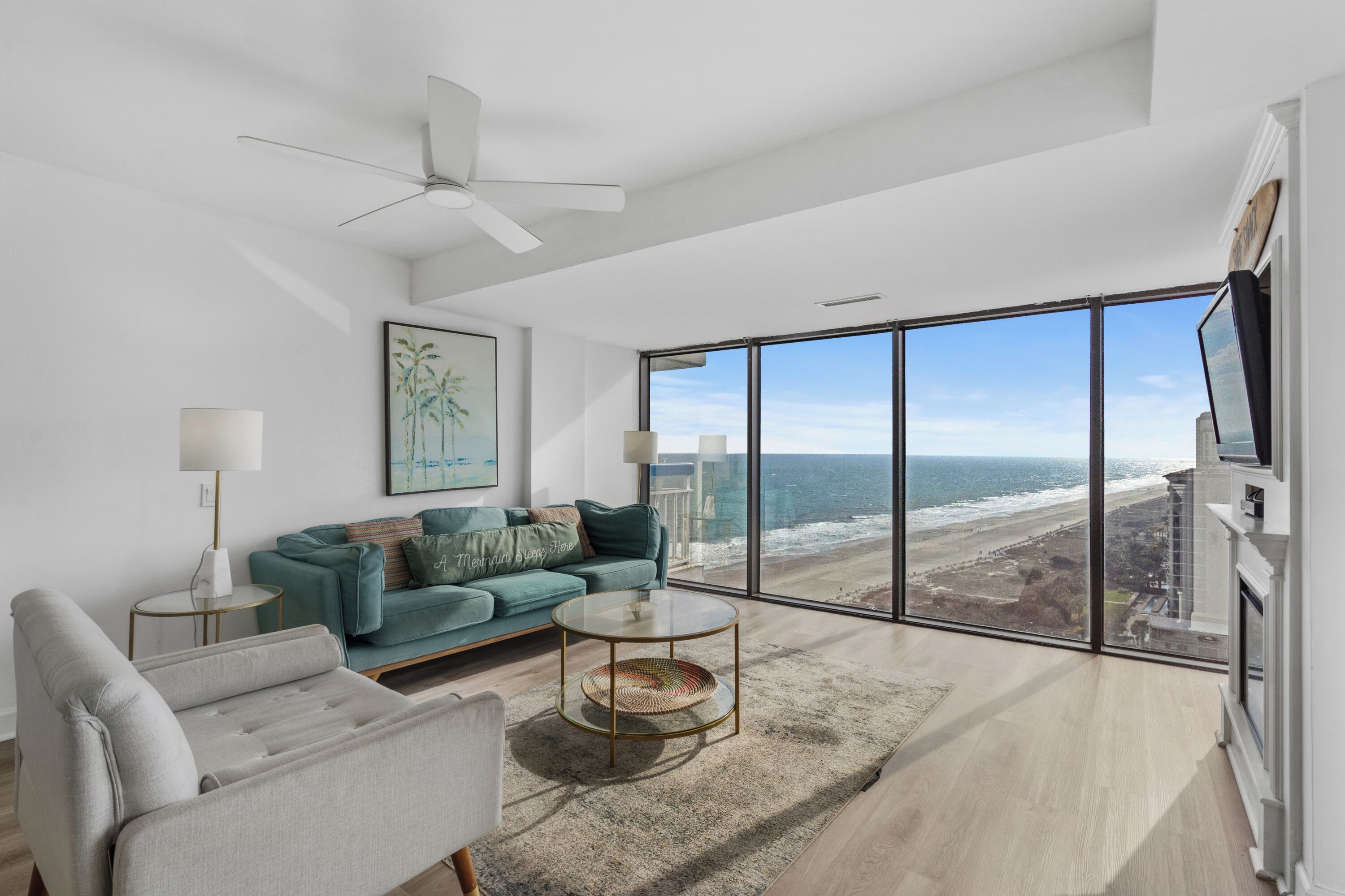 A modern living room with a large window overlooking the beach, a ceiling fan, and a cozy seating area with a sofa and armchairs.