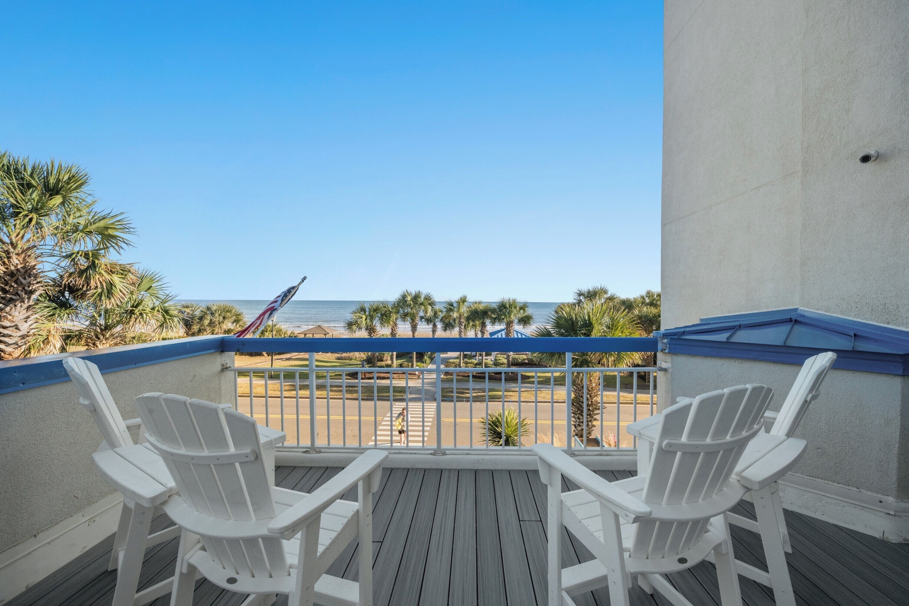 A balcony with two white Adirondack chairs, a view of the beach, and a palm tree.