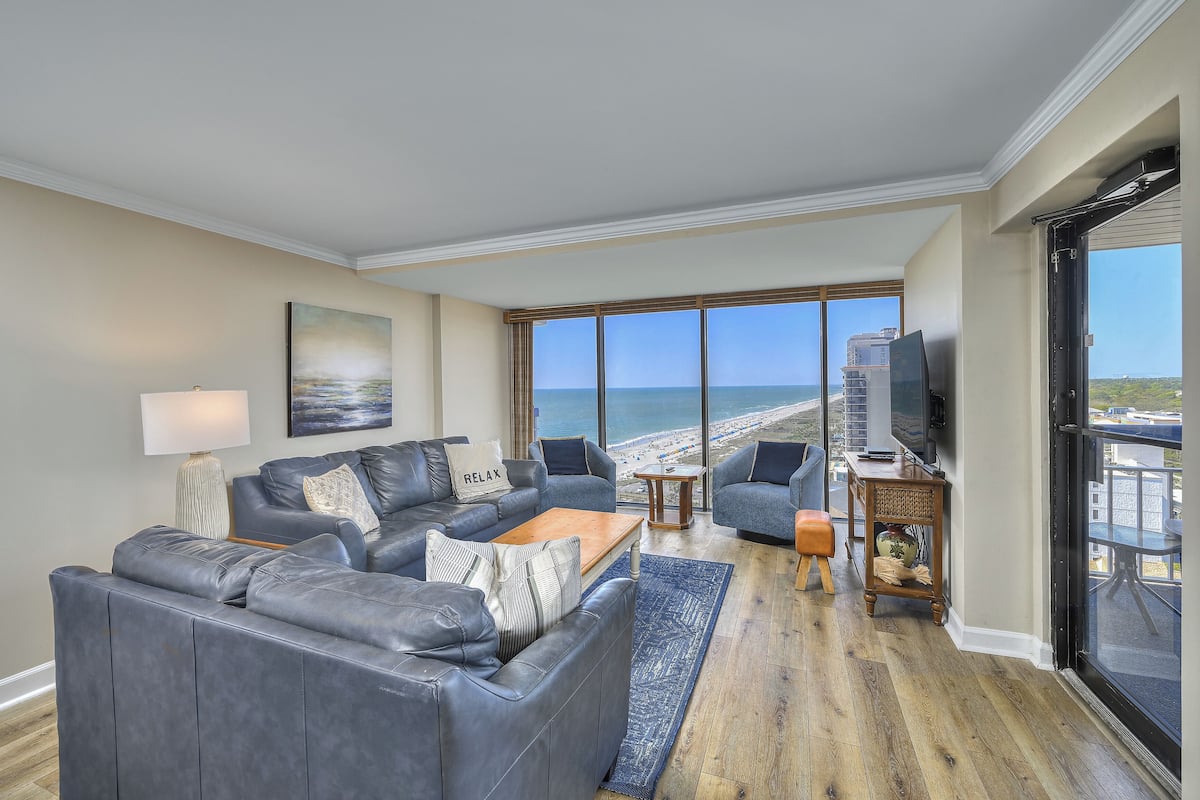 A living room with a large gray sectional sofa, a wooden coffee table, and a view of the beach through large windows.