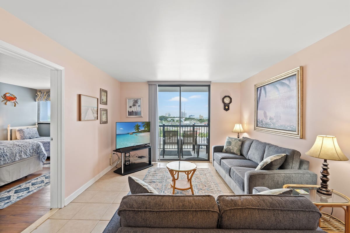 A living room with a sofa, armchair, and a TV. There is a balcony with a view of the city.