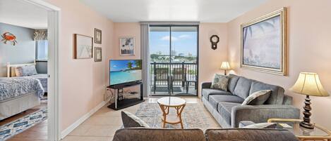 A living room with a sofa, armchair, and a TV. There is a balcony with a view of the city.