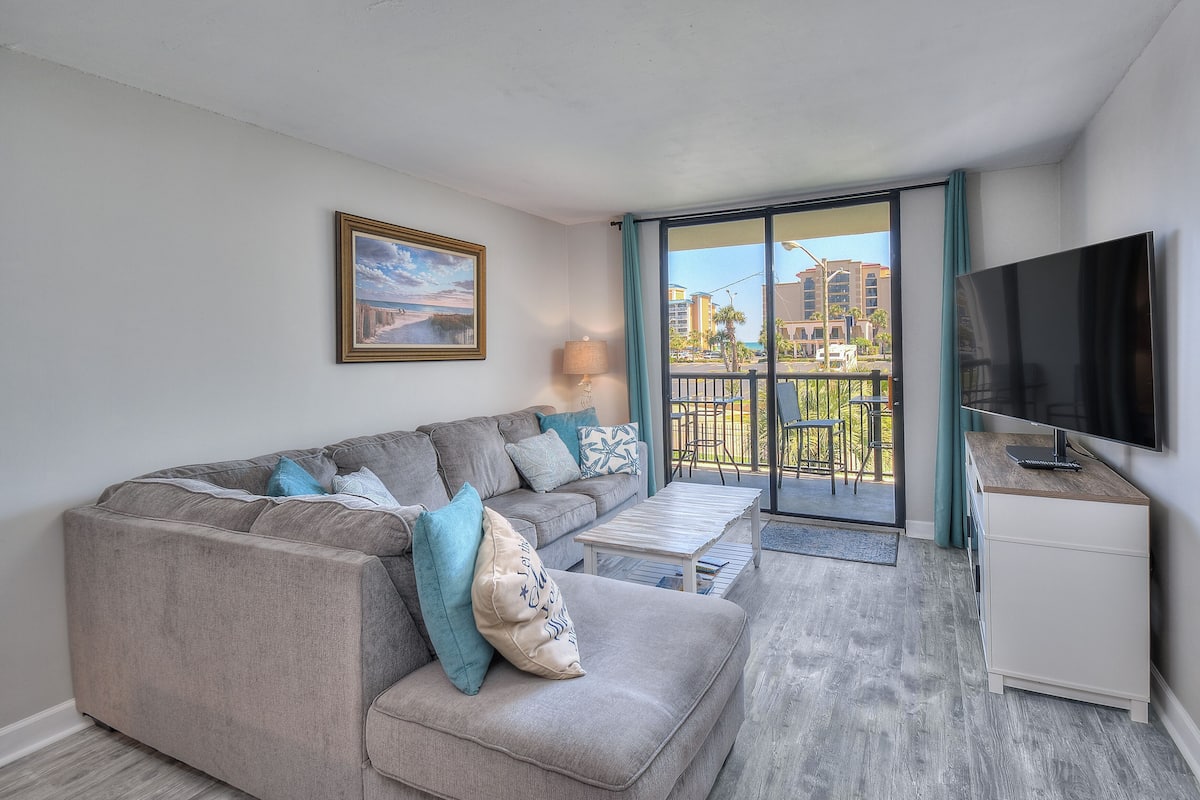 A modern living room with a grey sectional sofa, a flat-screen TV, and a balcony with a view of buildings.