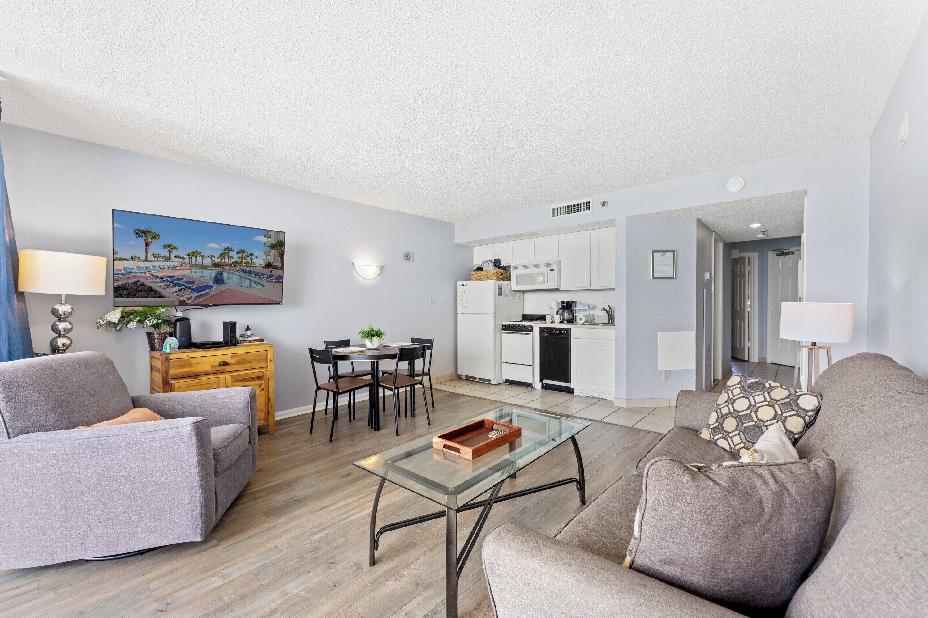 A modern living room with a glass coffee table, grey sofas, a dining area with a table and chairs, a kitchen with white cabinets, and a painting on the wall.