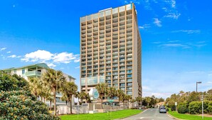 A tall, modern hotel with multiple floors and balconies, surrounded by lush greenery and palm trees. - Units at Ocean Forest Plaza by Elliott Beach Rentals (Myrtle Beach)