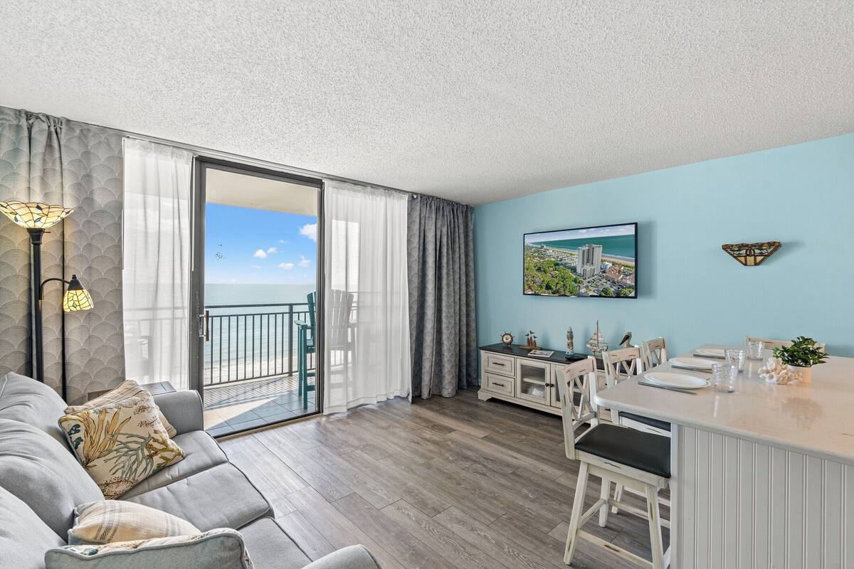 A modern living room with a sofa, dining area, and a large window offering a view of the sea.