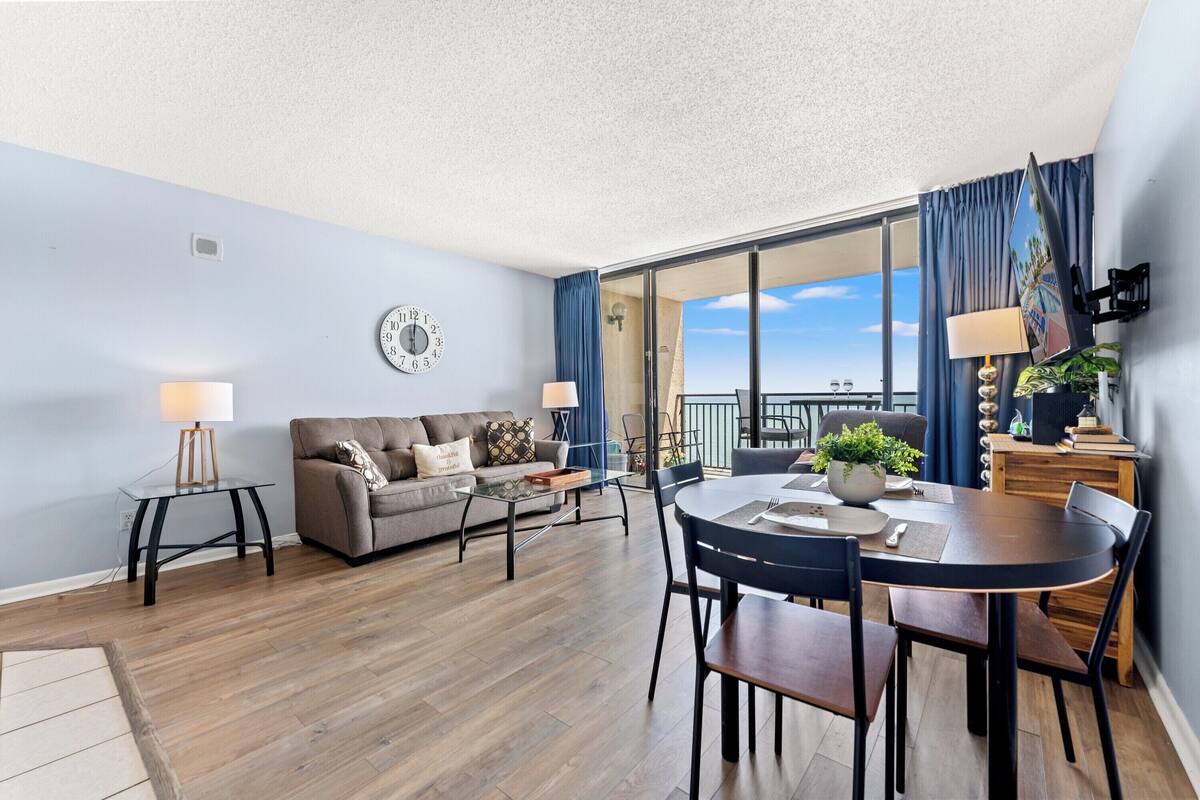 A modern living room with a sofa, coffee table, dining table, and chairs. There is a large window with a view of the sky and a balcony.