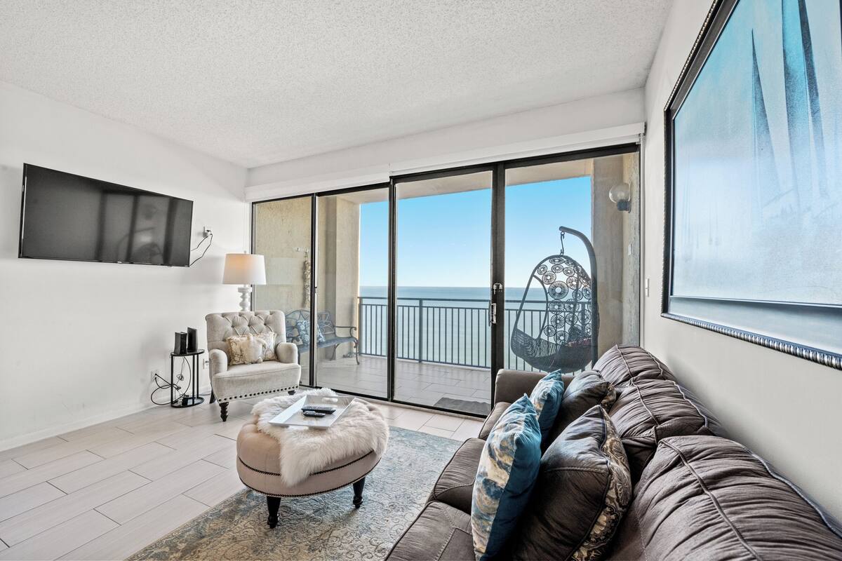 A modern living room with a sofa, ottoman, and armchair, a flat-screen TV, and a balcony with a view of the sea.