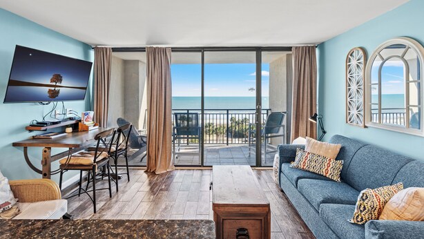 A living room with a sofa, TV, and dining area, overlooking a balcony with a view of the sea. - Units at Ocean Forest Plaza by Elliott Beach Rentals (Myrtle Beach)