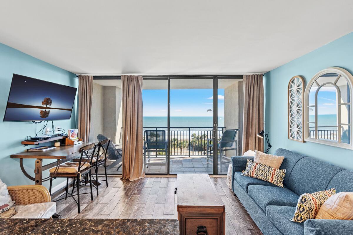 A living room with a sofa, TV, and dining area, overlooking a balcony with a view of the sea.