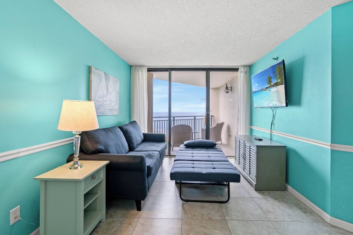 A room with teal walls, a sofa, a bench, a TV, and a view of the outdoors.
