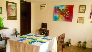 A dining room with a table covered by a white tablecloth and a colorful quilt. There are chairs around the table, a wooden cabinet, and a painting on the wall. - Casa Ceasars  Historic Quito (Quito)