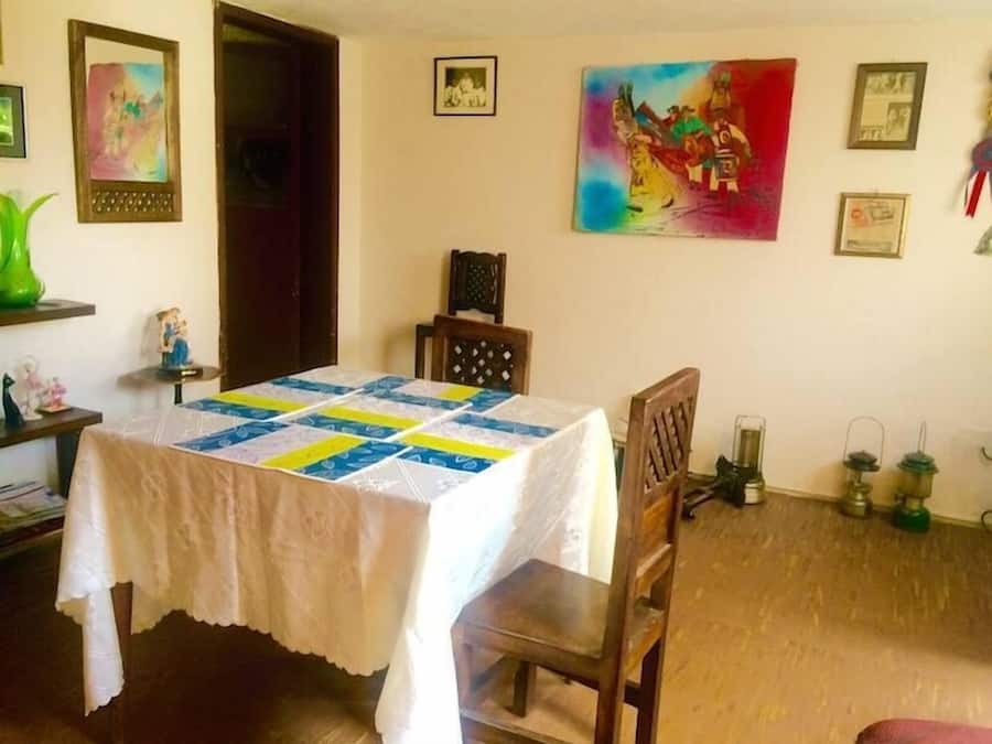 A dining room with a table covered by a white tablecloth and a colorful quilt. There are chairs around the table, a wooden cabinet, and a painting on the wall.