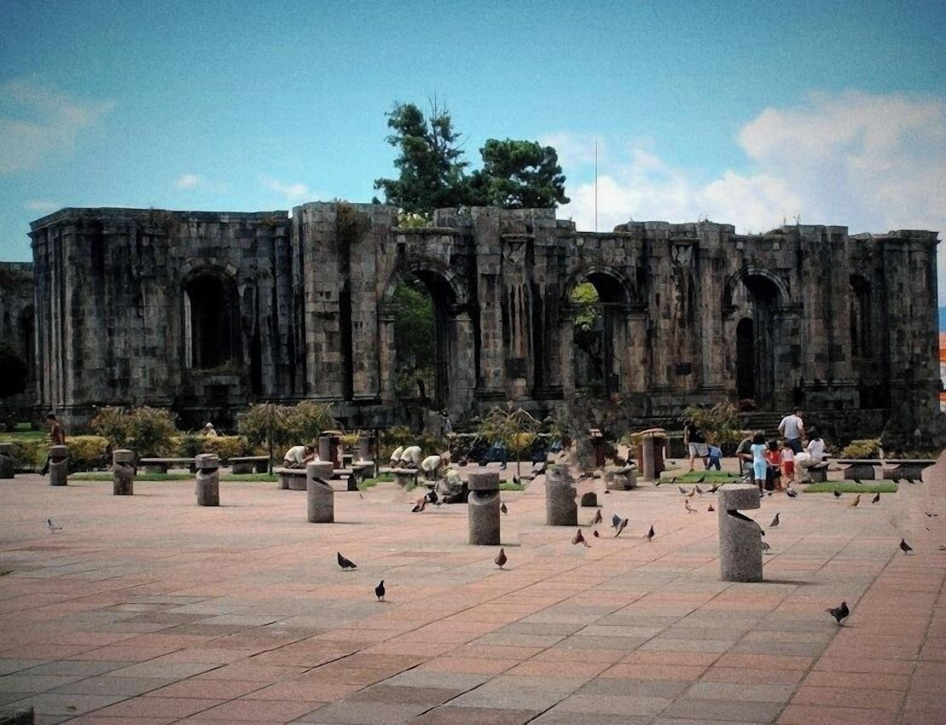 An ancient stone structure with multiple arches and columns, surrounded by a paved area with scattered birds.