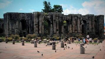 Una antigua estructura de piedra con múltiples arcos y columnas, rodeada por una zona pavimentada con aves dispersas.