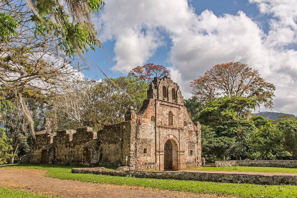 Un antiguo edificio de piedra con un arco central, rodeado de abundante vegetación y árboles.