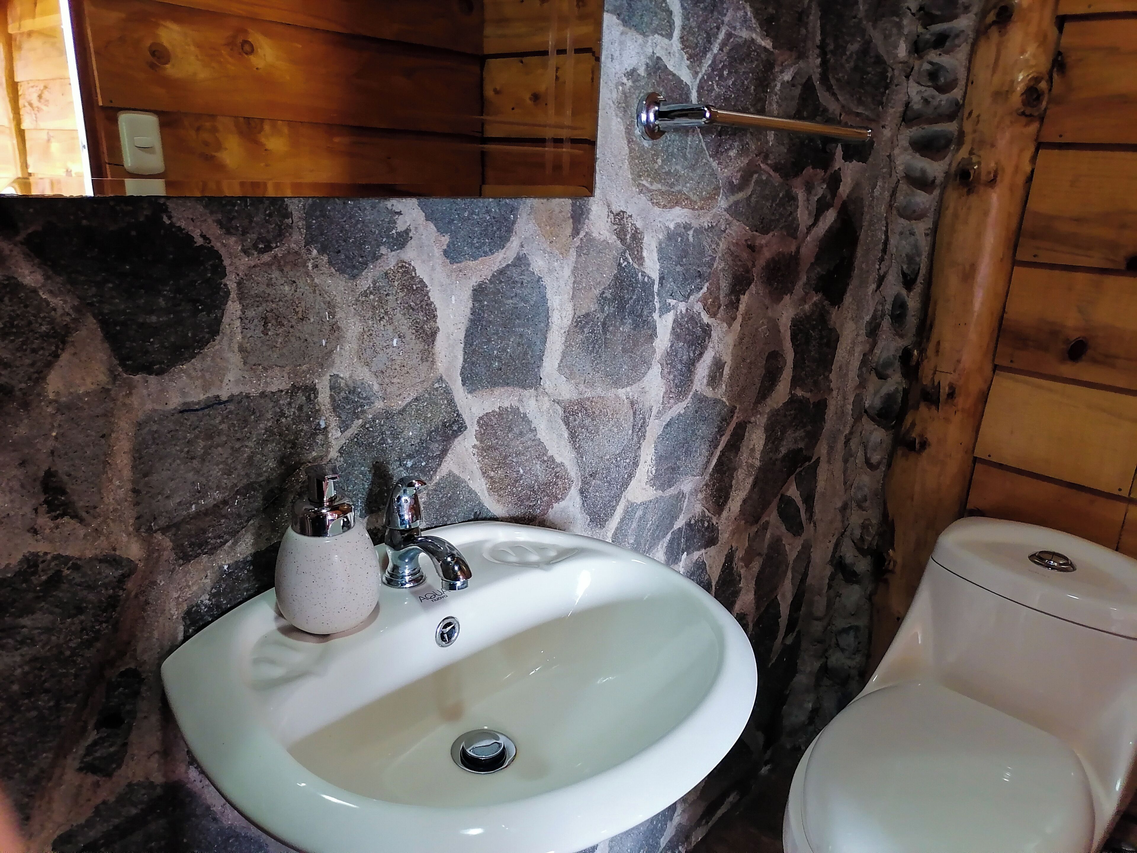 Une salle de bain avec un mur en pierre, un lavabo blanc doté de deux robinets et des toilettes.