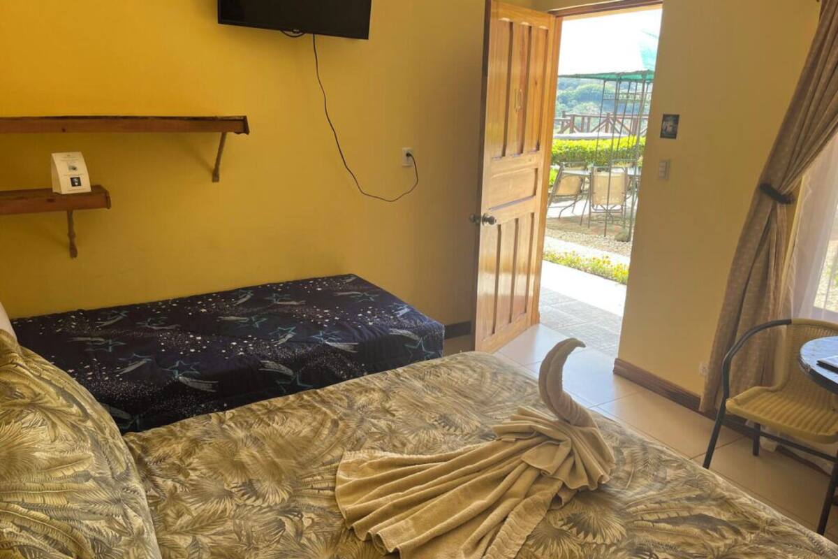 A room with a bed, a table, and a chair. There is a door leading outside.