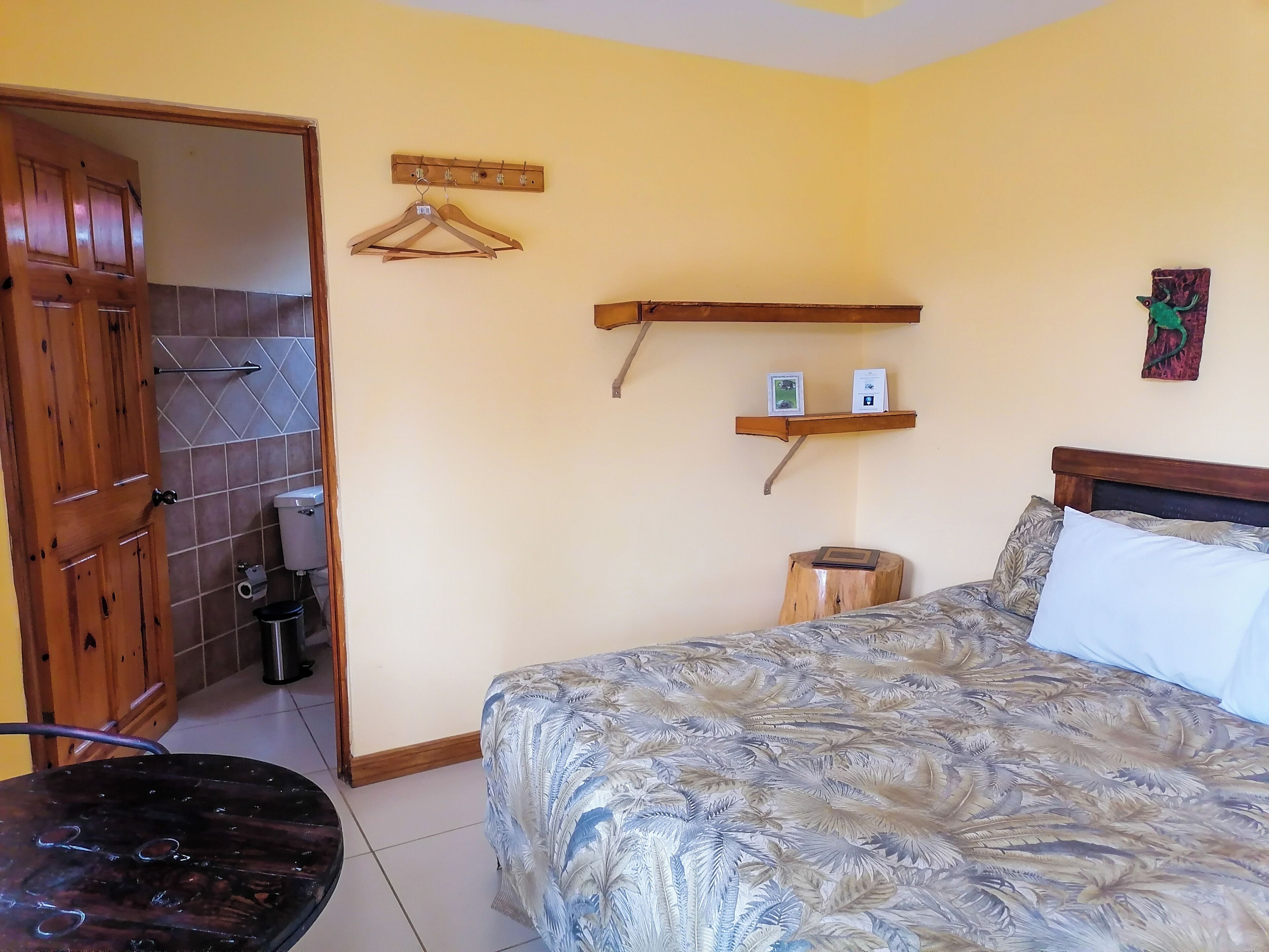 Une chambre à coucher avec un lit, une porte en bois, un petit tabouret, une étagère avec des objets et un porte-manteau fixé au mur.