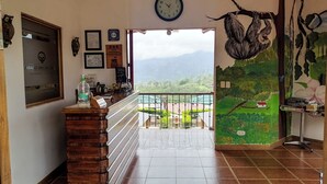 A reception area with a wooden counter, a clock, framed certificates, and a view of a mountainous landscape through an open door. - Paraíso Orocay Lodge (Paraiso)