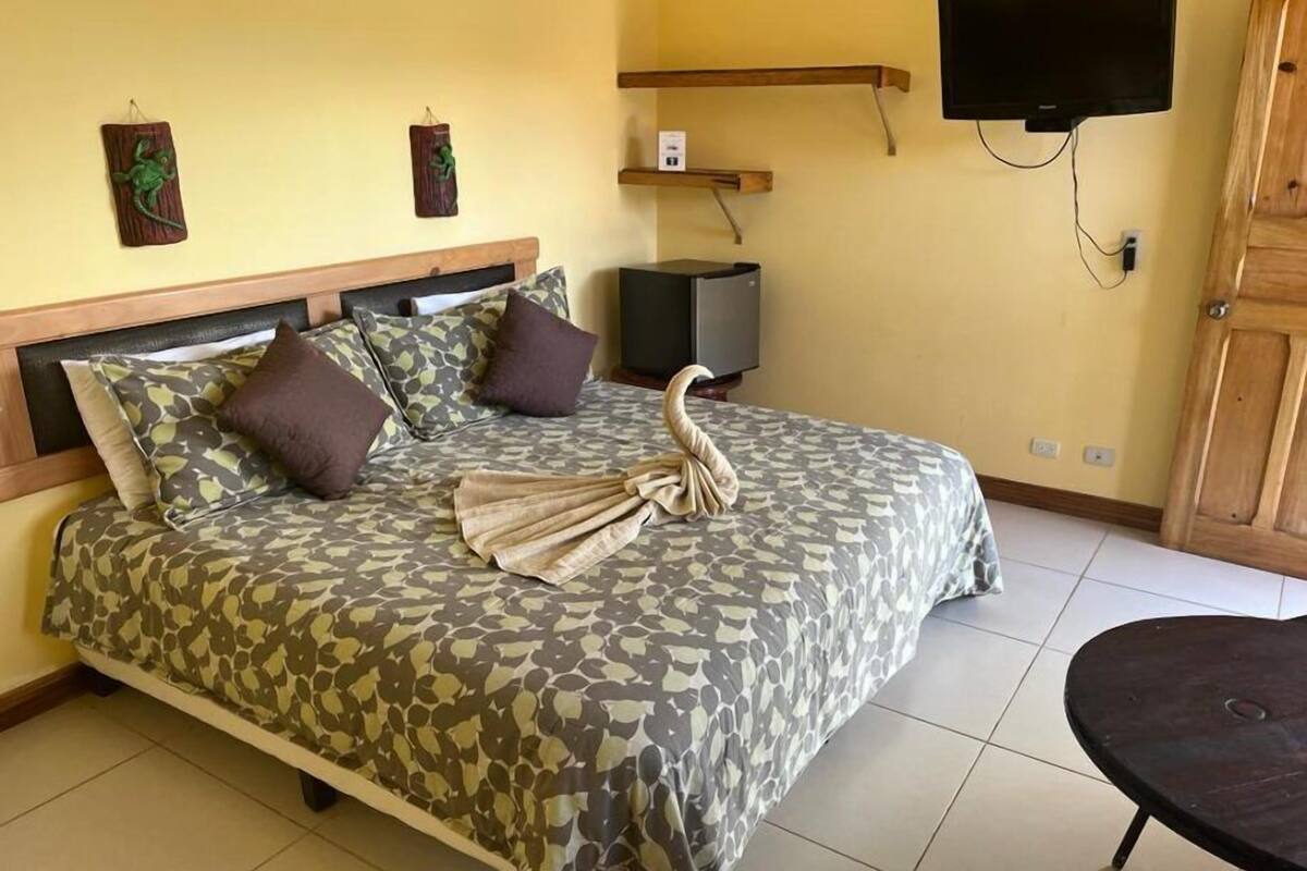 A hotel room with a bed, a flat-screen TV mounted on the wall, a wooden cabinet, and a small table.