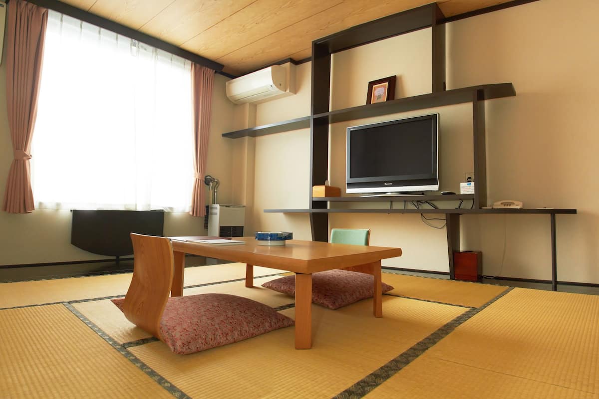 A room with tatami mats, a wooden table, two chairs, a television on a shelf, and a window with curtains.