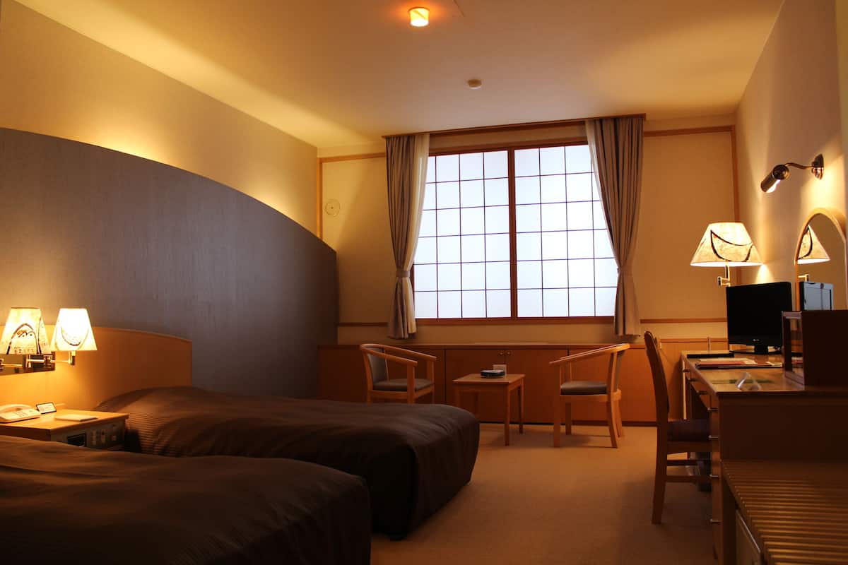 A hotel room with two beds, a desk, a chair, a TV, and a window with curtains.