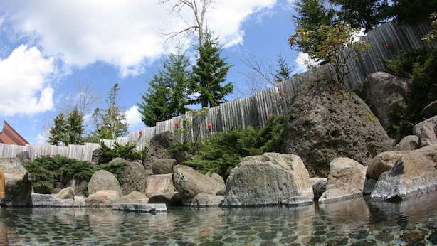 Una piscina cristalina rodeada de rocas grandes y árboles, con una valla de madera al fondo.