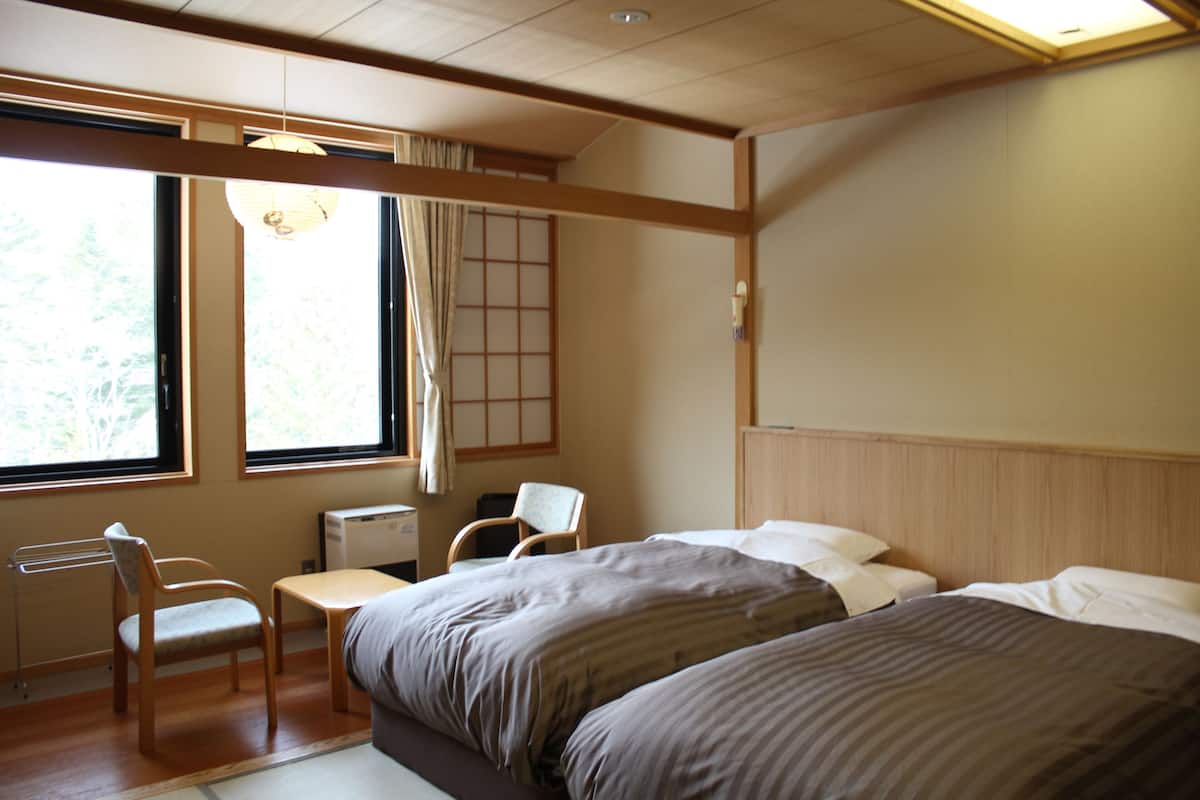 A hotel room with two beds, a desk, a chair, and a window with a view of trees.