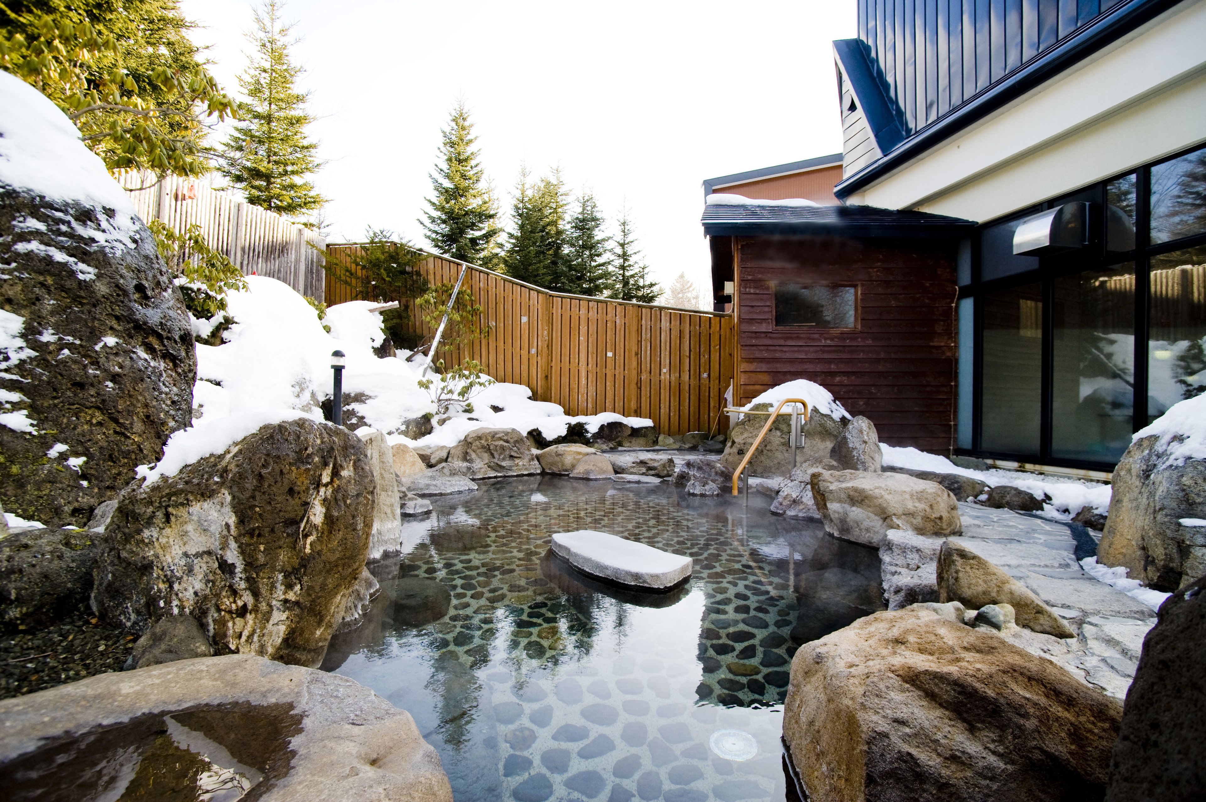 A hot spring surrounded by large rocks and snow, with a wooden fence and a modern building in the background.