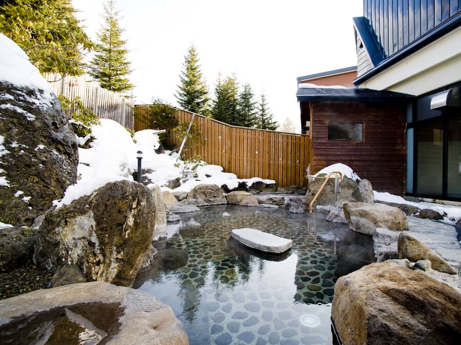 A hot spring surrounded by large rocks and snow, with a wooden fence and a modern building in the background.