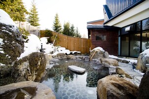 A hot spring surrounded by large rocks and snow, with a wooden fence and a modern building in the background.