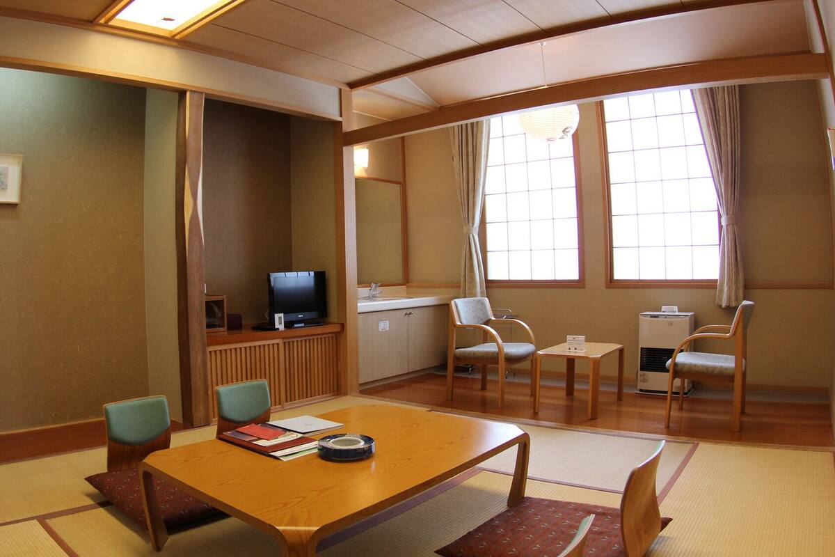 A traditional Japanese-style room with tatami flooring, a low wooden table, two chairs, a television, and a window with curtains.