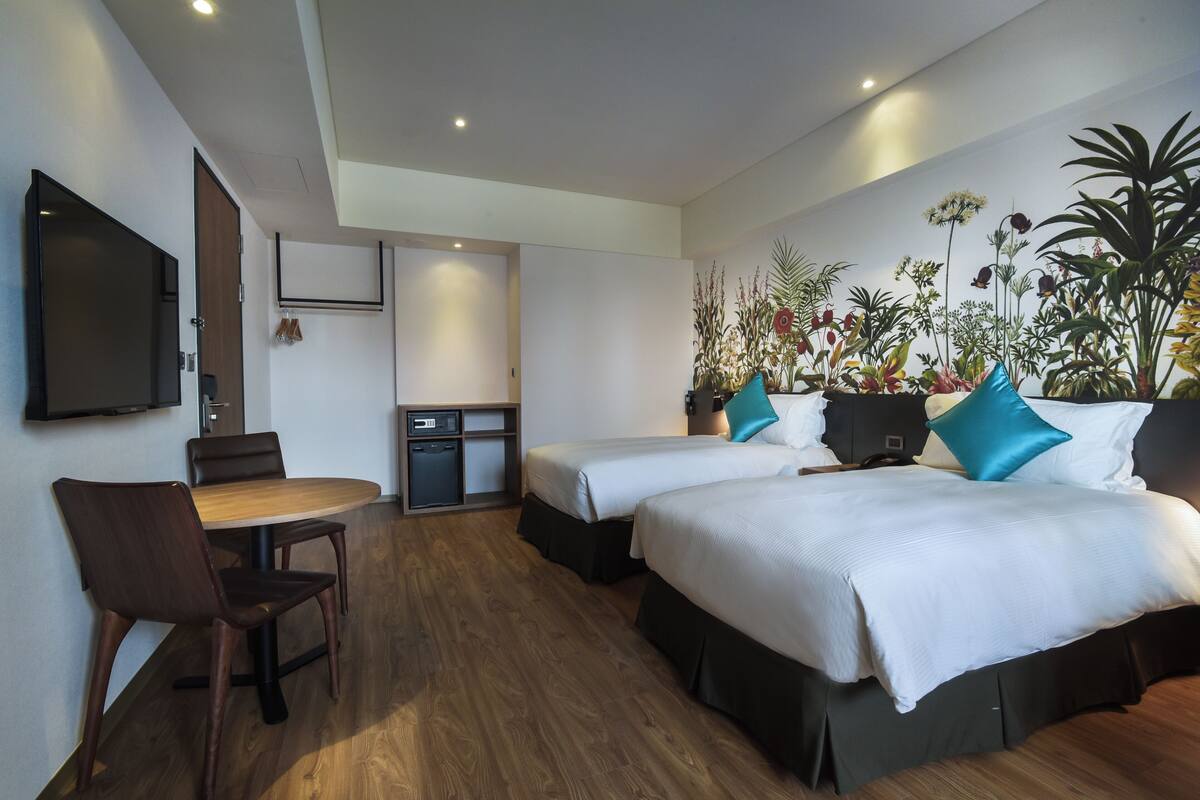 Habitación de hotel con dos camas, zona de comedor con mesa y sillas, televisor montado en la pared y una pequeña kitchenette.