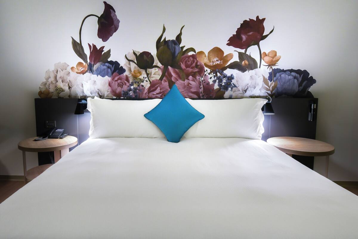 Habitación de hotel con una cama grande, un cojín azul decorativo y un mural floral en la pared.