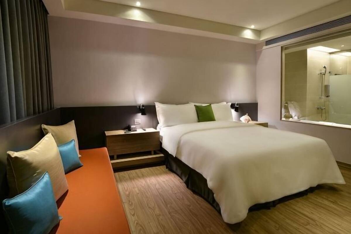 Una habitación de hotel con cama, mesitas de noche, un banco y un ventanal grande.