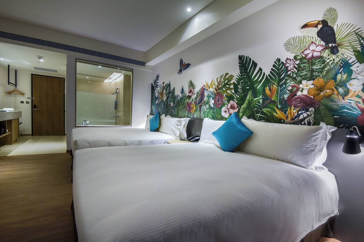 Habitación de hotel con dos camas, un espejo grande y un vibrante mural tropical en la pared.