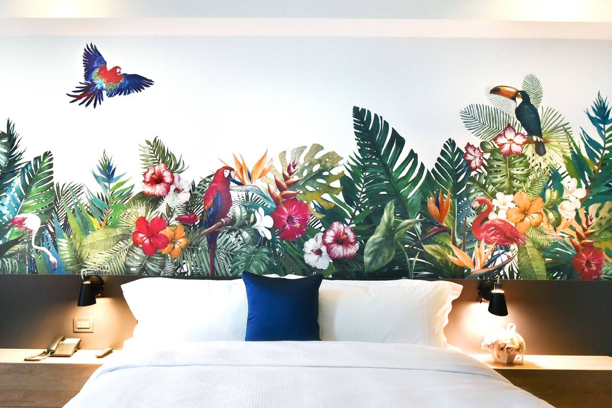 Una habitación de hotel con una cama grande, una mesita de noche con un teléfono y un mural en la pared que muestra plantas y aves tropicales.