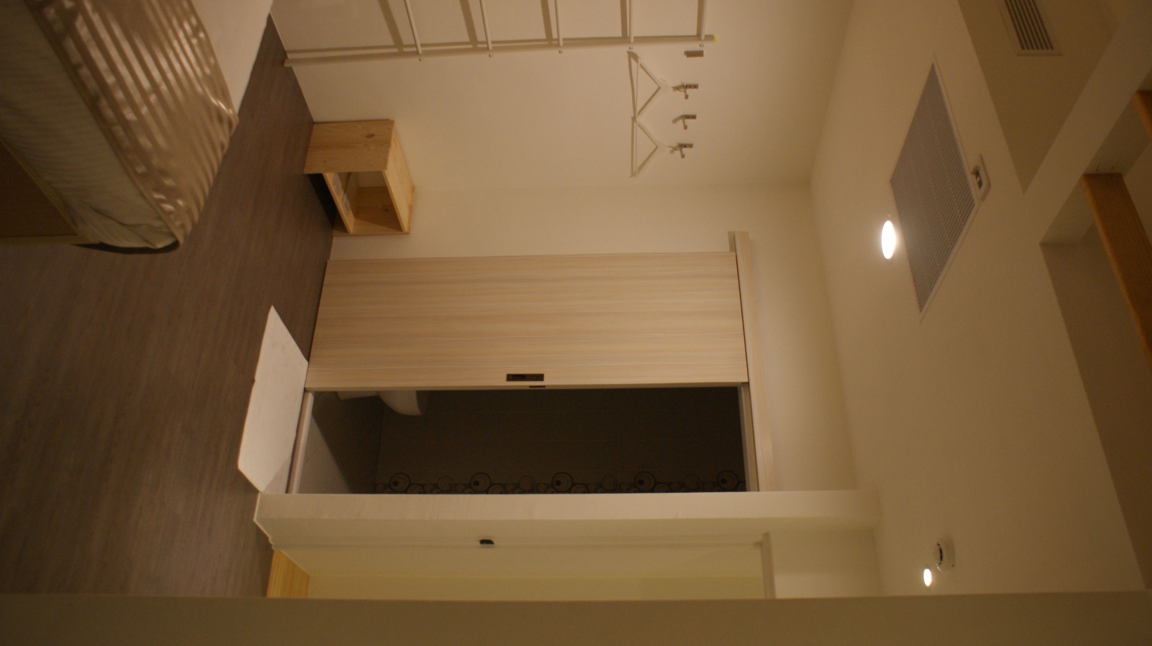 A room with a sloped ceiling, a wooden shelf, a window with blinds, and a vent on the wall.