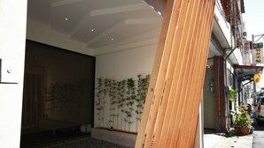 Un edificio moderno con una gran estructura de madera, una pared con líneas verticales y una calle con coches aparcados y plantas en macetas.