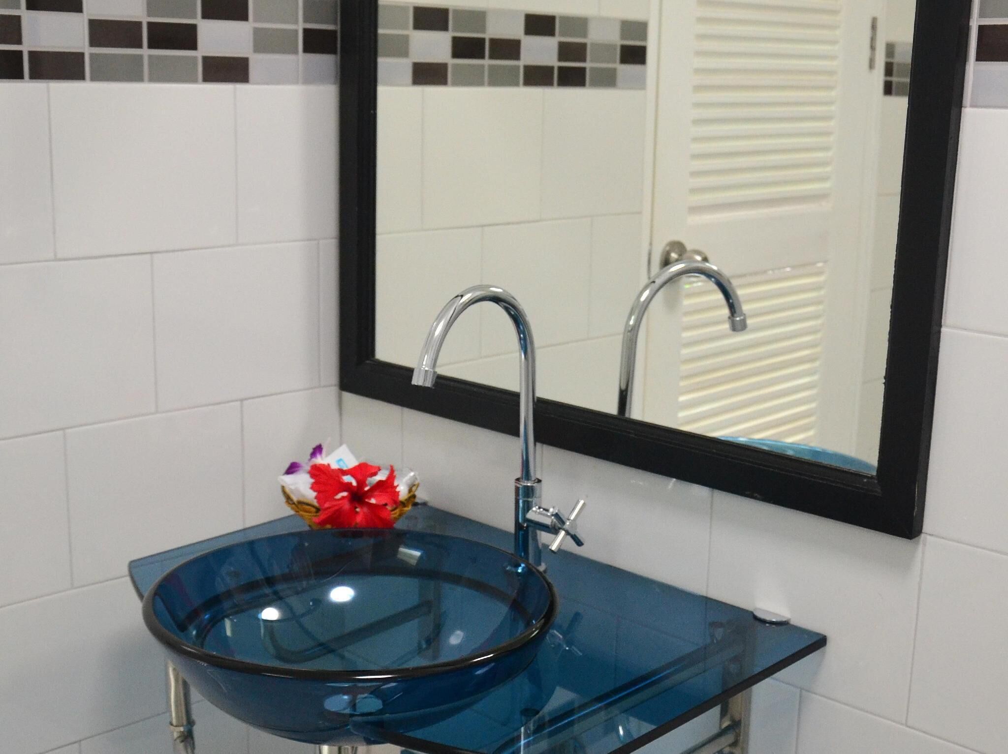 Un baño con un lavabo de vidrio, un espejo con marco negro y paredes con azulejos blancos.